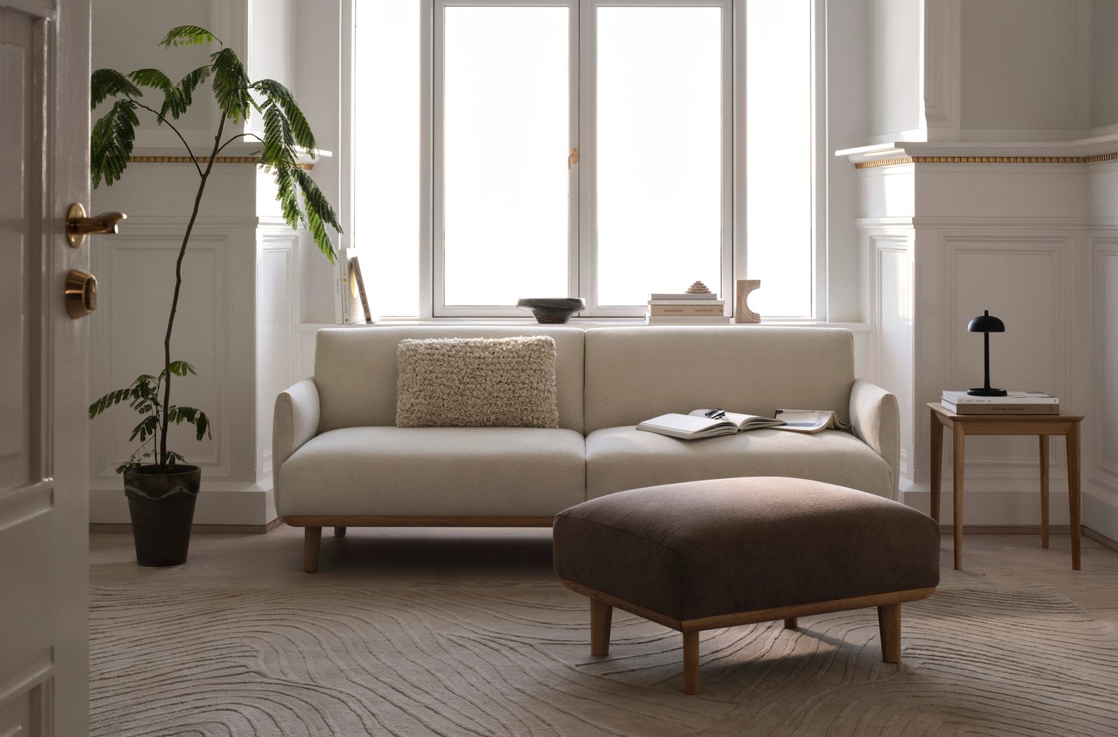 Helles Sofa mit gepolstertem Hocker vor einer Fensterfront, darunter ein gemusterter Teppich